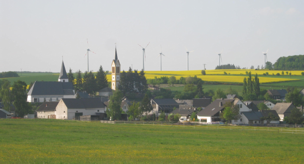 Kleiner Ort mit einer Kirche in einem grünen Feld umgeben von Bäumen und Pflanzen, Windmühlen im Hintergrund unter einem klaren blauen Himmel.