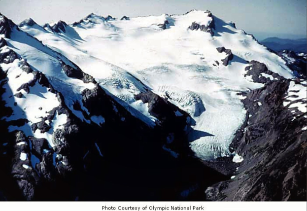 Majestätischer Berg mit einem großen Gletscher, umgeben von schneebedeckten Gipfeln und einem klaren blauen Himmel, mit Text am unteren Rand.