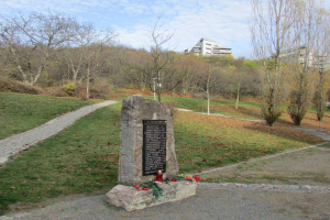 Ein Denkmal in einem Park mit einer Tafel, auf der "Gedenkstätte für die Opfer der Tschernobyl-Katastrophe" steht, umgeben von Gras, trockenen Blättern, einem Weg, Bäumen, Pflanzen, einem Straßenmast, Gebäuden und einem bewölkten Himmel.